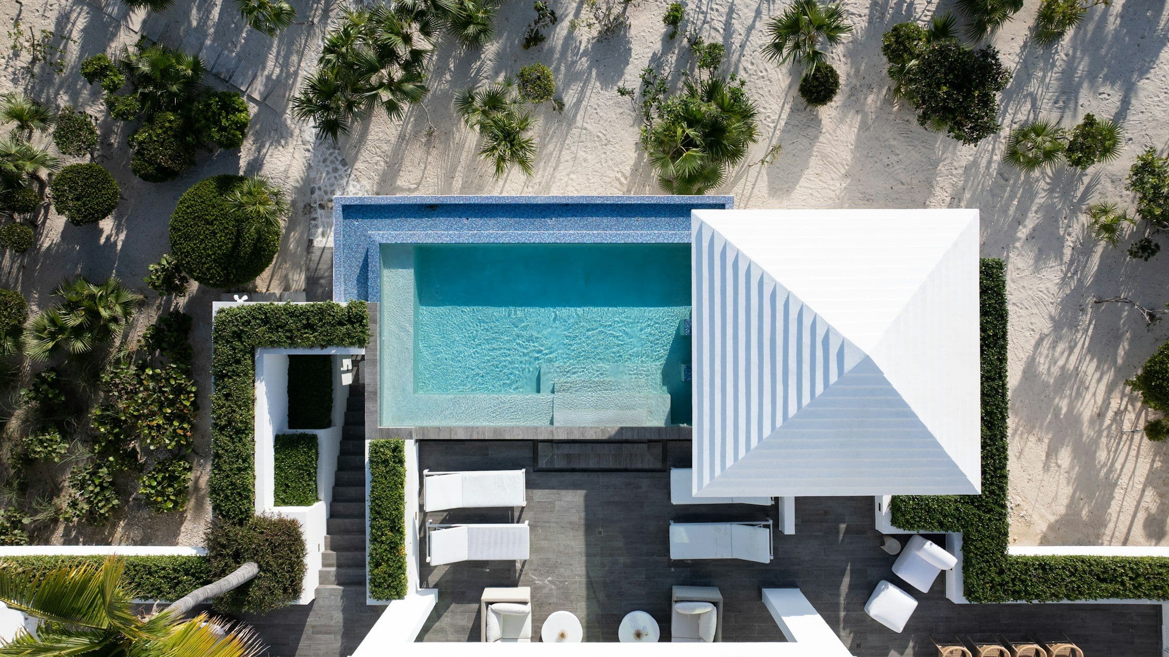 Overhead view of a white roof, pool deck and loungers among palms