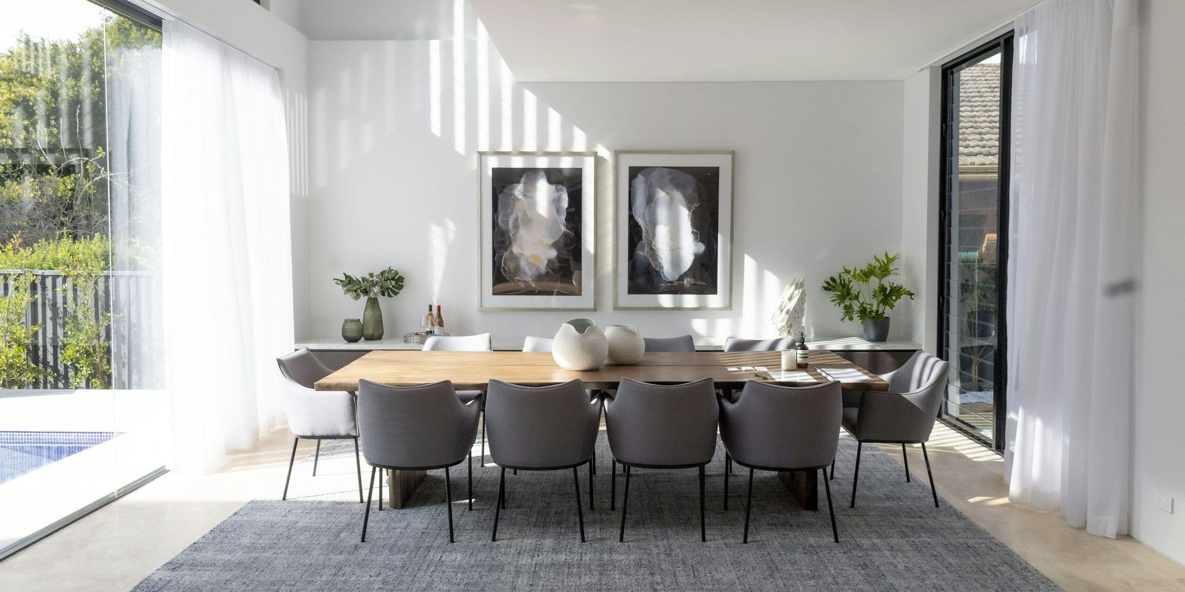 White dining room with a long table and a pool beyond the glass doors