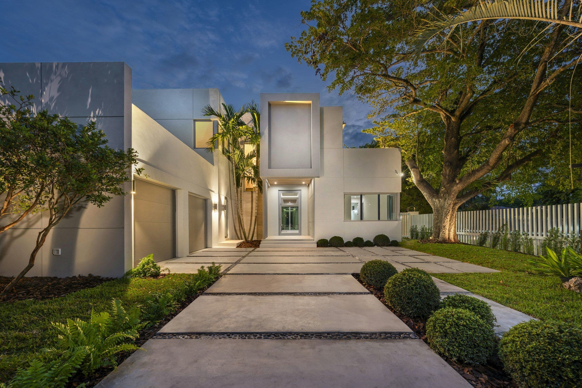 White contemporary home with a lit glass entry and palms at twilight